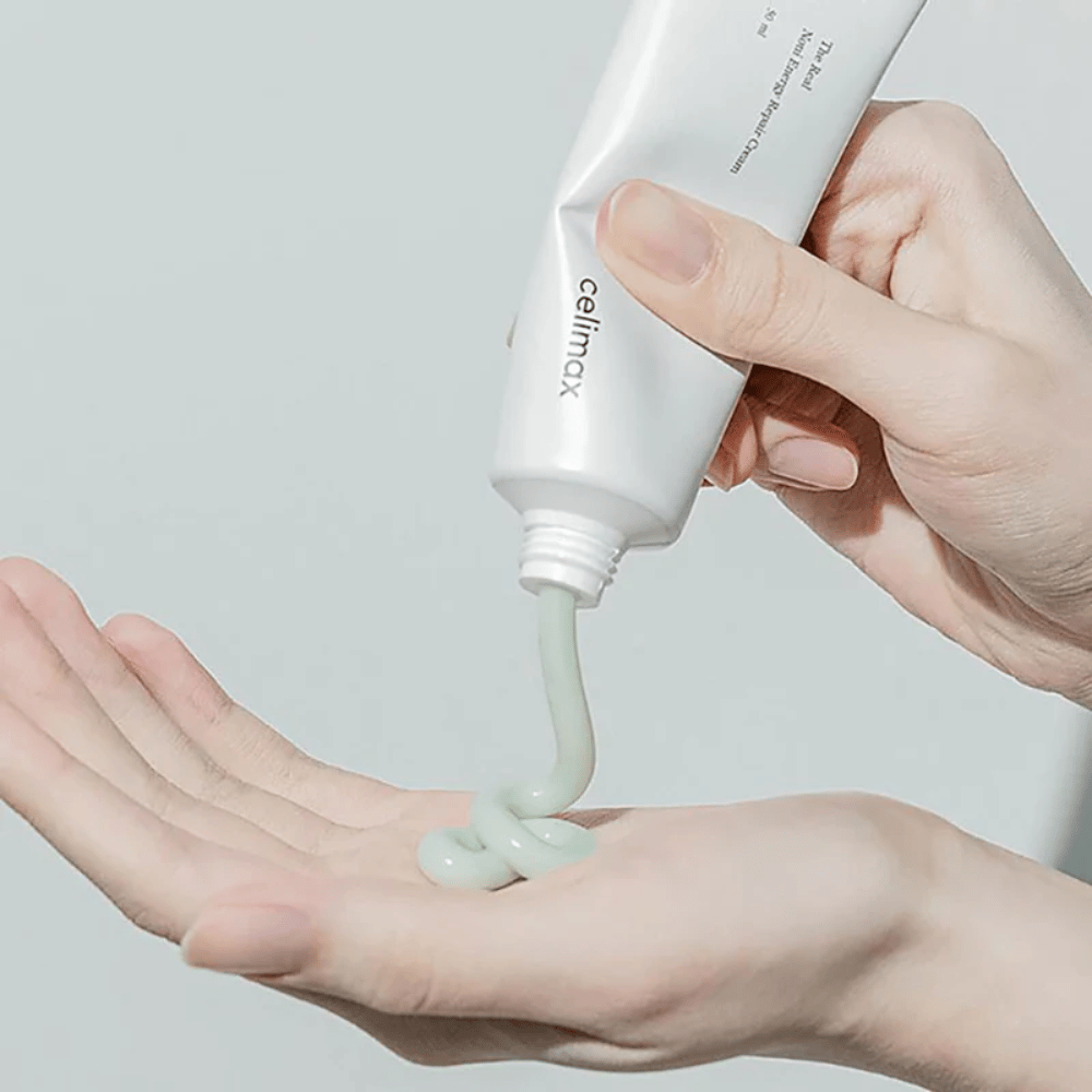 A hand holds a white tube of Celimax Noni Energy Repair Cream, and the cream is being squeezed out in a smooth stream onto the palm. The cream is light green and marketed to nourish and restore the skin.