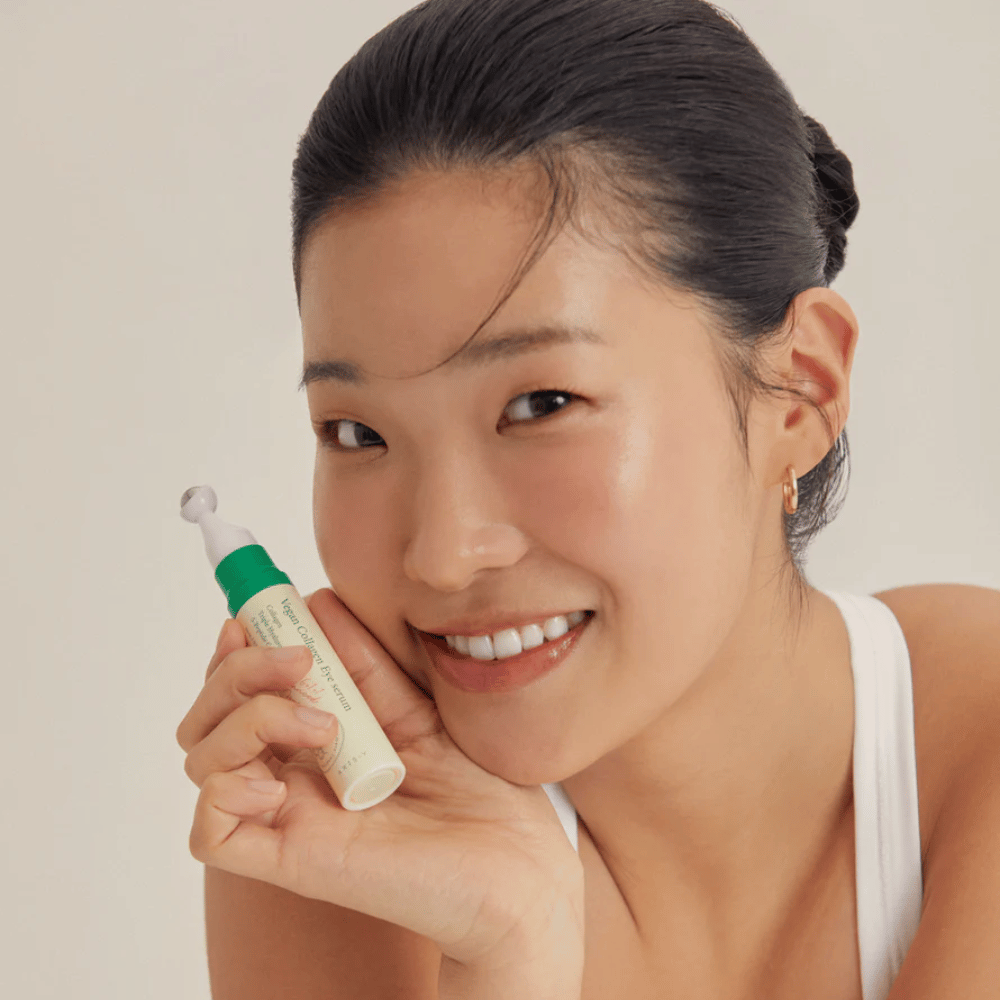 A woman with her hair up and golden earrings holds a AXIS-Y Vegan Collagen Eye Serum 10ml close to her face, while she smiles softly. The serum is designed to moisturize and reduce wrinkles around the eyes, specially formulated for sensitive skin.