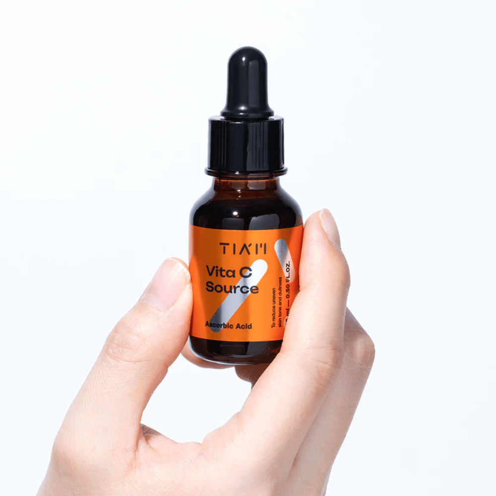 Hand holding TIA'M Vita C Source, a vitamin C serum in a brown glass bottle with a pipette. The product is designed to even out skin tone and add radiance.