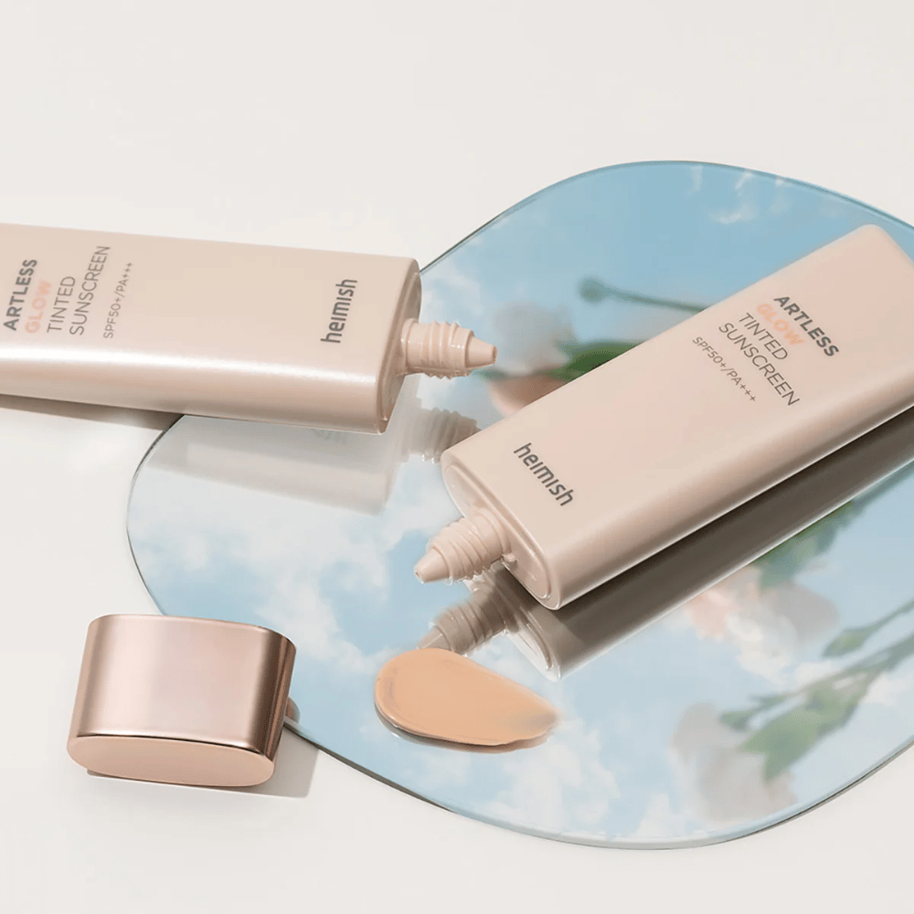 A bottle of heimish Artless Glow Tinted Sunscreen SPF50+ PA+++ 40ml and a bottle of concealer on a mirror, showing different skin tones.
