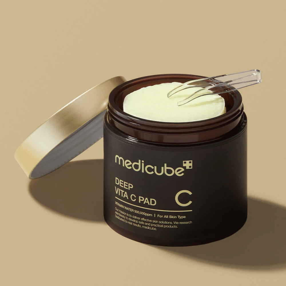 A jar with medicube Deep Vita C Pad. The jar is dark brown with gold text and a white, inverted label with vitamin C information. The inside of the lid is gold, and a transparent tweezer lies on top of the light yellow cotton pads inside. The background is solid beige.