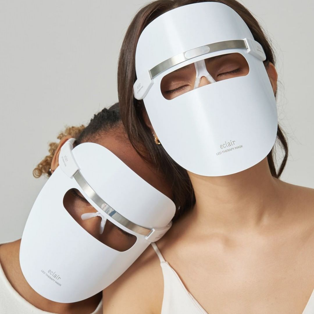 Two women wearing white masks demonstrate ECLAIR LED Therapy Mask for skin tone improvements.