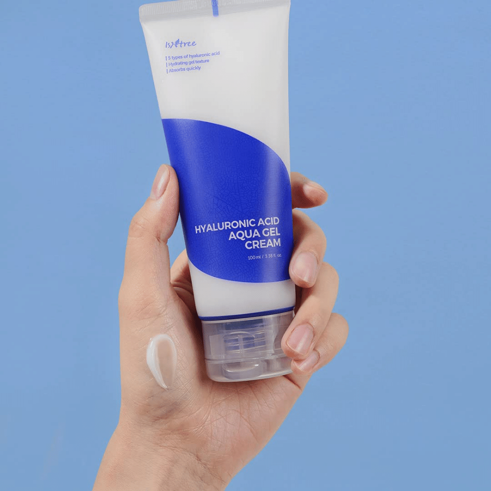 A hand holding a tube Isntree Hyaluronic Acid Aqua Gel Cream 100ml on a blue background.